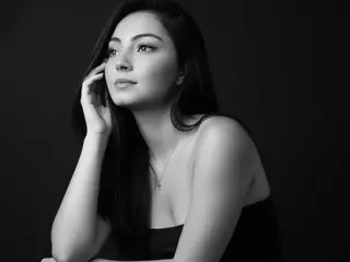 ViolletaRosse poses with her hand to her face, showing her long dark hair and bare shoulders against a black background.