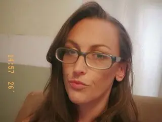 Brunette woman with glasses looking thoughtfully at the camera in a cozy indoor setting.