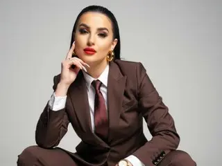 Elegant woman in a brown professional suit and red lipstick posing confidently against a grey background.