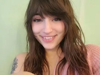 VixenMinx smiles with long brown hair, wearing a pink top against a light green background.