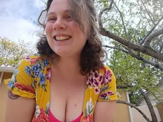 WendiHo smiles while wearing a bright yellow floral print top that shows off her cleavage, with her wavy hair framing her face against a leafy tree background.