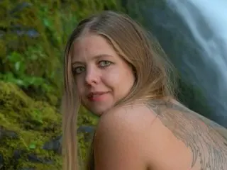 Blonde woman with a large wing tattoo posing in a lush green mossy forest setting.