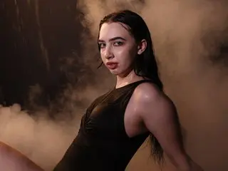 WildKittenX poses with long dark hair, wearing a black sleeveless top against a dark, smoky background.