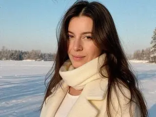 XandraK poses with long dark hair wearing a white scarf and cream coat against a snowy outdoor background.