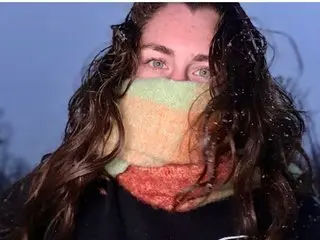 Xintrist wears a thick, multi-colored knit scarf wrapped around her face, showing only her eyes and long, wavy dark hair against a blue sky.