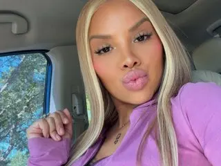 XkayluvX poses with long blonde hair and pink lipstick, wearing a purple top while sitting in a car.