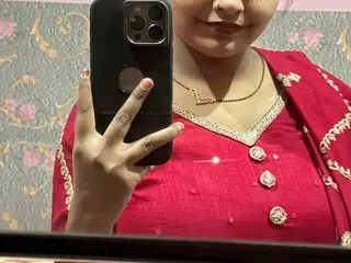 YOURMISTHI poses with a smile, wearing a red patterned top with white trim, while holding a black smartphone in front of her face.