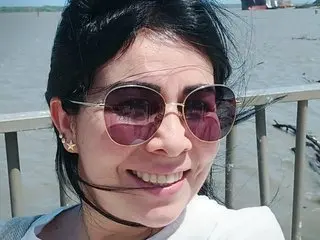 Yanella_109 poses with short black hair and sunglasses, wearing a white top against a soft outdoor background.