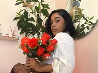 YariRose poses with a bunch of red roses, wearing a white shirt and black lace stockings, with green plants in the background.