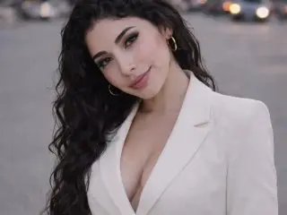 YennettSphinx poses with long, dark wavy hair, wearing a white blazer with a deep cleavage showing her chest.