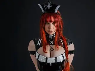 YourWhiteTulip wears a black and white cosplay outfit with a headpiece, showing cleavage and her long red hair against a dark background.
