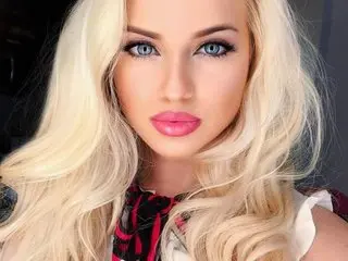 Beautiful blonde woman with blue eyes and pink lipstick wearing a patterned top in a soft, glamorous portrait.