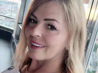 Smiling blonde woman in casual attire enjoying a view from a high-rise city window.