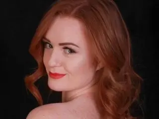 Zara_Nevada_xx poses with her long red hair down, looking at the camera with a smile against a black background.