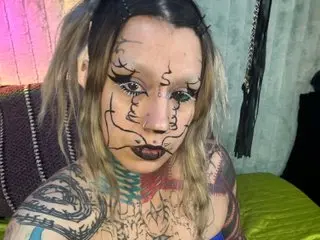 Zaturn shows her face and chest, featuring heavy black facial tattoos and dark lipstick, while posing in a dimly lit room.