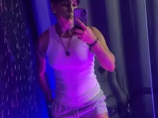 ZhoeSexyGirl poses in a white tank top, showing her toned arms and neck against a dark, purple-lit background.