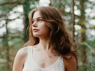 ZlataDiamonds poses with her long brown hair down, wearing a white lace top against a soft, natural outdoor background.