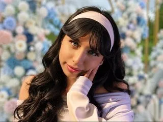 ZoeBell with long wavy dark hair and a white headband poses against a background of blue and white flowers.