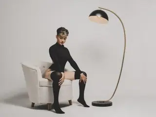 Fashionable woman in a black turtleneck and high socks sitting on a white armchair by a modern floor lamp.