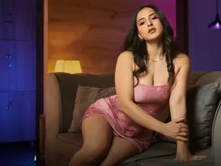 ZoeLambert poses in a pink satin slip dress, leaning against a dark sofa with her long dark hair down and a direct gaze.