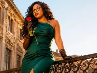 ZoeQueens poses on a balcony wearing a green satin dress and holding a red rose.