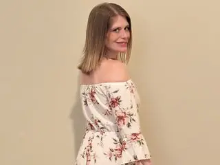 ZoeeB poses sideways, looking back over her shoulder with her long blonde hair down, wearing a white off-the-shoulder floral print top.