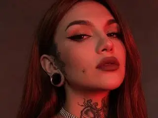 Zoenina69 poses with a direct gaze, showing her long red hair, dark eye makeup, and a small mole on her chin and cheek, with a visible ear piercing.
