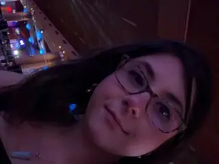 adrilately looks toward the camera with a soft smile, wearing glasses and letting her dark hair fall over her shoulders.