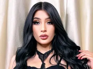 Glamorous woman with long wavy black hair and dramatic makeup wearing a black lace outfit against beige curtains.