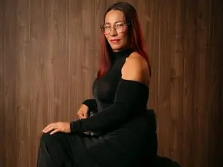 agatha_fit is a woman with long dark hair and glasses wearing a black long-sleeved dress, posing against a wooden wall.