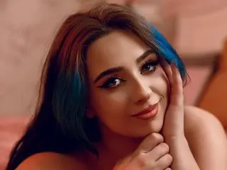 Smiling young woman with blue highlighted hair posing in a cozy, warm-lit bedroom setting.