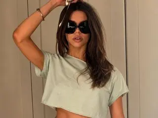 ameliawinx poses with her hands behind her head, wearing a light green t-shirt and black sunglasses with her long brown hair down.