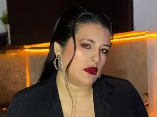 andreasecret poses with a direct gaze, wearing a black blazer and silver hoop earrings, with her dark hair pulled back.