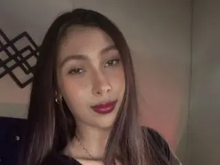 angeldreams21 looks at the camera with long dark hair and red lipstick, wearing a dark top against a plain background.