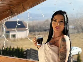 arianatattoos poses with long dark hair, wearing a white top and showing her chest tattoos while looking at the camera outdoors.