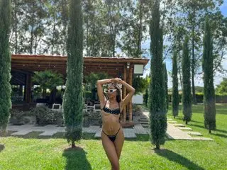 Slim woman in black bikini posing in a sunny garden with tall cypress trees and a wooden pergola.