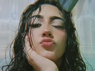 babbyvalen poses with a pouty lip, showing her long dark wavy hair and eyes looking down in a close-up shot.