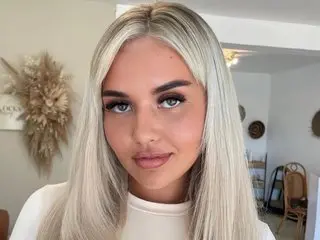 babypeachy poses with her long blonde hair down, wearing a white top against a neutral indoor background.