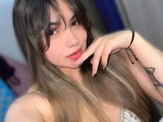Young woman with long brown hair and bangs wearing a white lace top in a soft, dreamy indoor setting.