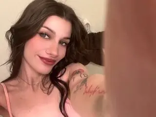 Smiling brunette woman in a pink camisole with tattoos posing for a selfie in a bright indoor setting.
