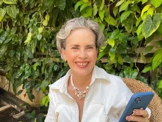belleblondmature smiles while wearing a white button-up shirt, showing her blonde hair and teeth against a background of green leaves.