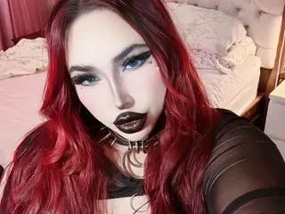belledarkgoddess with long red hair and dark lipstick, wearing a black choker and sheer black top, looking directly at the camera.