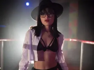 betty_hot wears a black bra and white open shirt, posing with long dark hair and glasses in a dim studio setting.