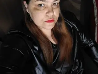 boommbi poses with long brown hair, wearing a black shiny leather jacket against a dark background.