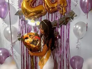 briithanyAdams99 poses in a white outfit with long dark hair, surrounded by gold balloons and purple streamers in a decorated room.