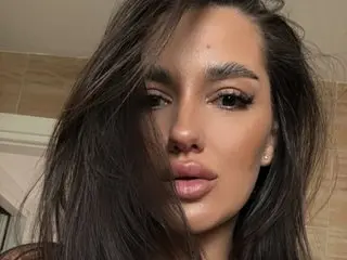 Close-up portrait of a beautiful woman with long brown hair and glamorous makeup in a warm indoor setting.