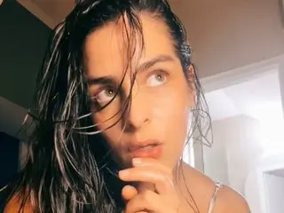 Thoughtful young woman with wet hair in a soft-lit indoor setting wearing a white strap top.