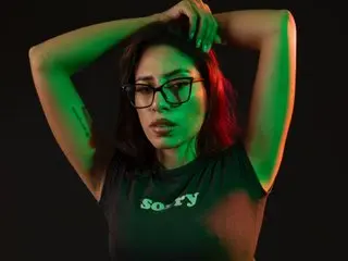 carla_palmer poses with her hands behind her head, wearing a black t-shirt and glasses against a dark background.