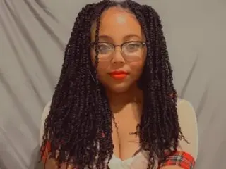 carmelloveskisses has long dark braids and wears a white top while posing against a grey background.
