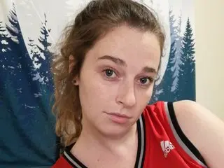 ceceeee looks at the camera with a neutral expression, wearing a red tank top with white trim, with her light brown hair down.